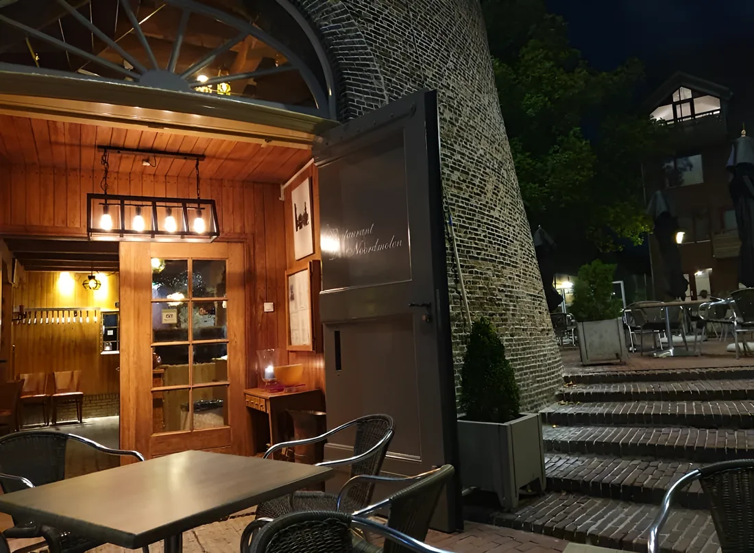 De verlichte ingang van het restaurant onder de boog van de molen, met terrastafels op de kade en de trap naar de kade