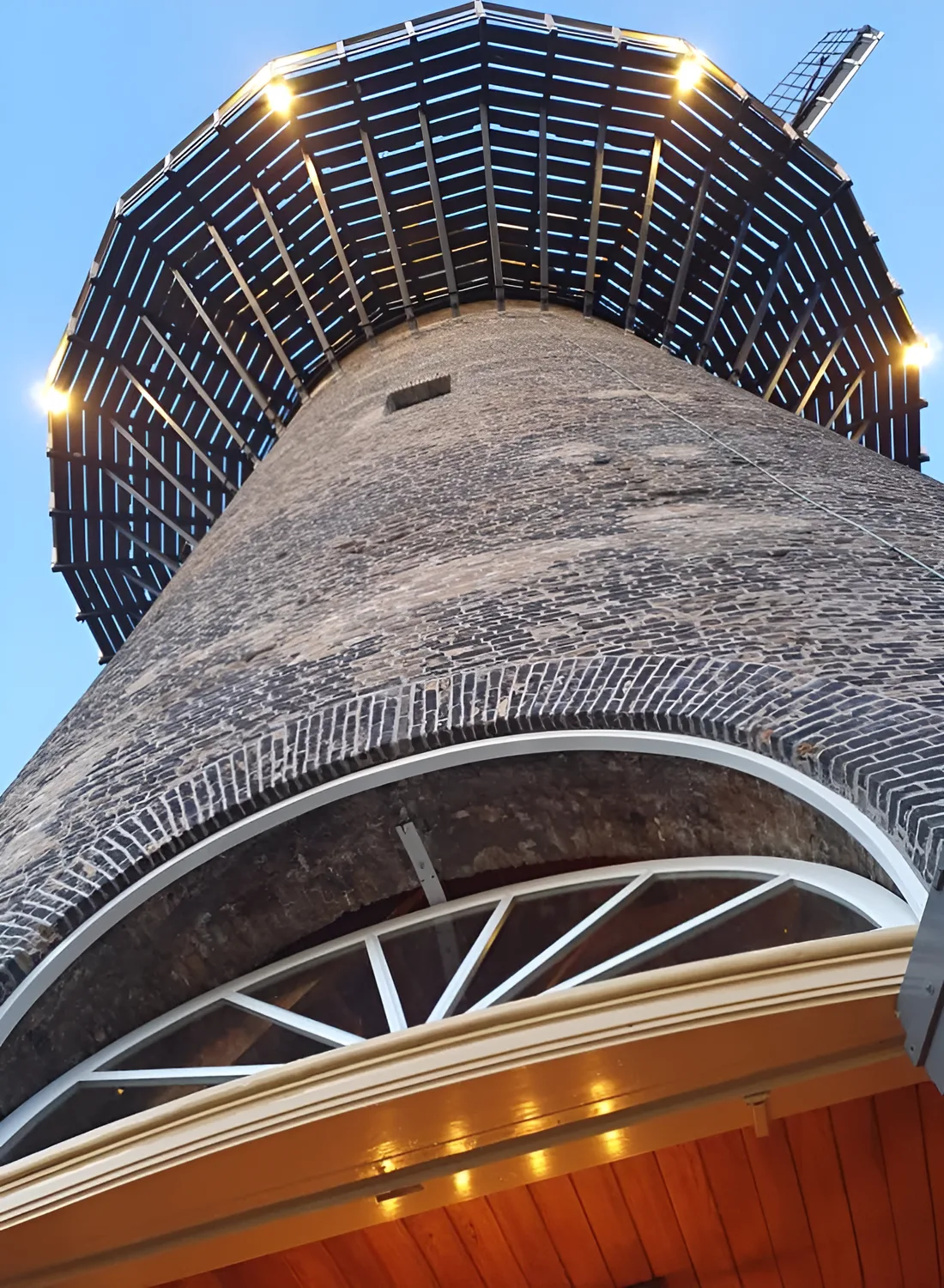 Van beneden af omhoog kijkend langs het metselwerk van molen De Noord naar de verlichte balie en de wieken